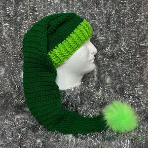 May include: A handmade green elf hat with a long, pointed end and a bright green pom-pom. The hat features a dark green body and a lighter green band around the forehead. The hat is made of a soft, knitted material.