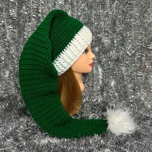May include: A green and white crochet elf hat with a long, pointed shape and a fluffy white pom-pom at the end. The hat is made of green yarn with a white band at the base. The hat is displayed on a mannequin head.
