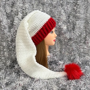 May include: A handmade crochet hat with a long, pointed tail. The hat features a white body, a red band, and a red pom-pom at the end of the tail. The hat is designed to be worn during the winter season.