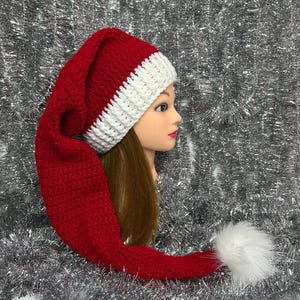 May include: A handmade red and white crochet Santa hat with a long, pointed end and a white pom-pom. The hat is displayed on a mannequin head with long brown hair, against a silver tinsel background. The hat is a festive accessory.