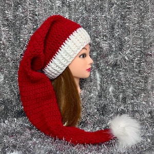 May include: A long, red crocheted hat with a white ribbed brim and a white faux fur pom-pom at the tip. The hat features a deep red yarn construction with a thick, textured white band around the base, designed in a classic elongated stocking style.