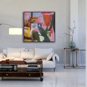 May include: Abstract painting with pink, green, yellow, and blue colors. The painting is framed and hanging on a white wall in a living room setting.