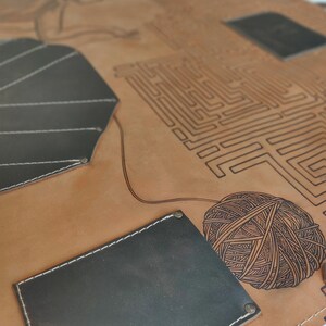 May include: A brown leather apron with a black and white maze design and a black leather pocket. The apron also features a black and white yarn ball design.