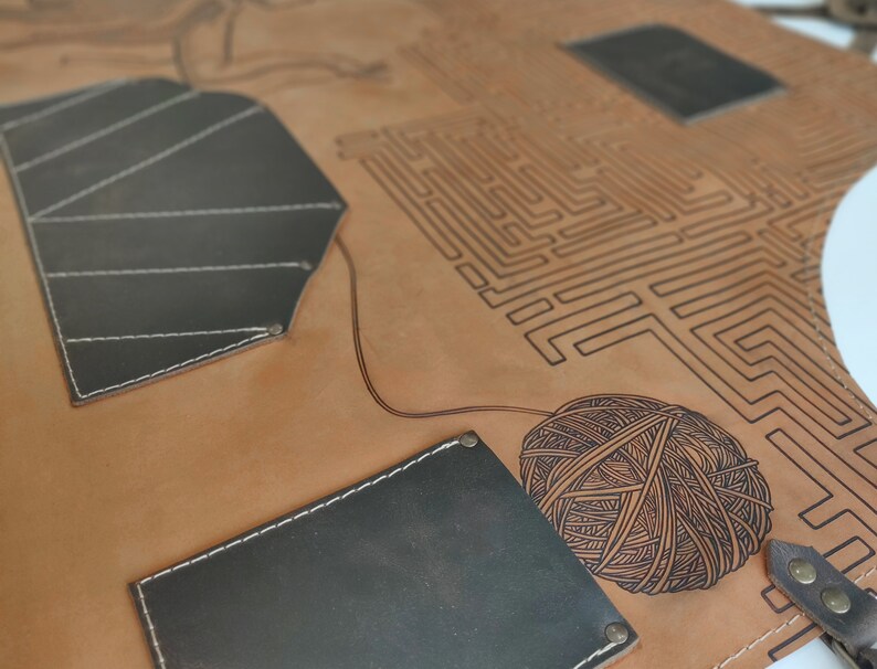 May include: A brown leather apron with a black and white maze design and a black leather pocket. The apron also features a black and white yarn ball design.