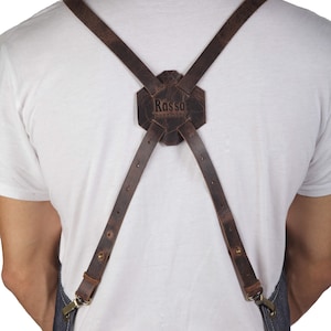 May include: A brown leather apron with adjustable straps and a buckle closure. The straps have a  "Rasso handmade" logo on the back.