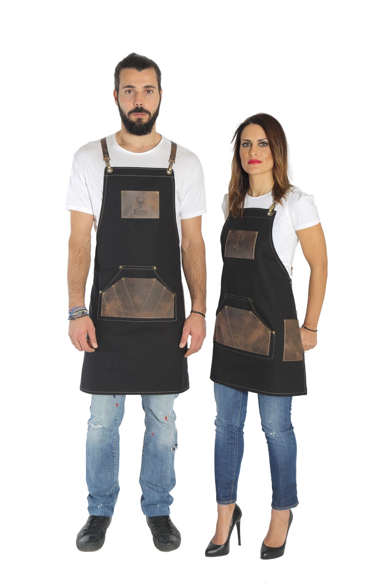 May include: Two people wearing black aprons with brown leather accents and the word "Rooster" on the front. The aprons have a large pocket on the front. The people are wearing jeans and white t-shirts.