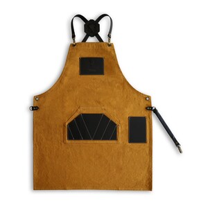 May include: A mustard yellow canvas apron with black leather straps, a large black leather pocket, and a smaller black leather pocket on the right side. The apron has a small black leather patch with the word "Maker" on it.