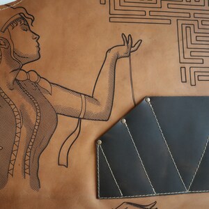 May include: A brown leather apron with a black leather tool pouch. The apron features a line drawing of a woman with a maze in the background.