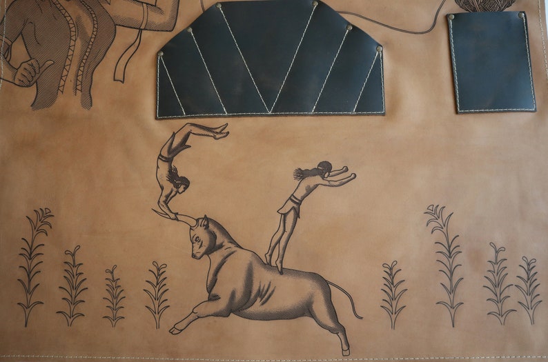 May include: A brown leather apron with a black leather patch and two smaller black leather pockets. The apron has a design of a bull, a woman jumping over the bull, and another woman doing a backflip.