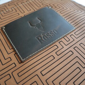 May include: A brown leather apron with a black leather patch featuring a bull head logo and the word "R&ouml;sso" embossed in the patch. The apron has a geometric pattern of black lines.
