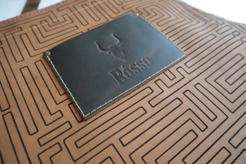 May include: A brown leather apron with a black leather patch featuring a bull head logo and the word "R&ouml;sso" embossed in the patch. The apron has a geometric pattern of black lines.