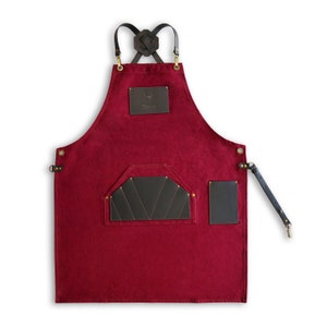 May include: A burgundy canvas apron with brown leather straps and accents. The apron has a large pocket with a brown leather flap and a smaller pocket on the right side. The apron has a brass buckle and the word "Dasso" is printed on a leather patch.