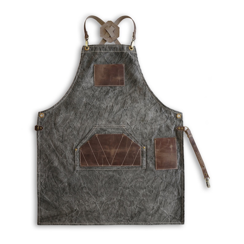 May include: A gray canvas apron with brown leather straps and pockets. The apron has a criss-cross strap design on the back and a large pocket on the front.