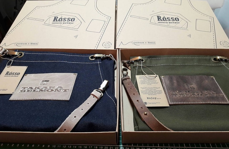 May include: Two canvas aprons with leather straps and a brown leather patch with the text "Takara Belmont" on each. The aprons are in a navy blue and a dark green color. The aprons are in boxes with the text "R&aacute;sso" on the top.