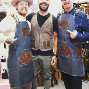 May include: Three people wearing denim aprons with leather accents. The aprons have a large pocket on the front. The person on the left is wearing a cowboy hat. The person in the middle is wearing a brown vest. The person on the right is wearing a black beret and glasses. The text "Rasso" is visible in the background.