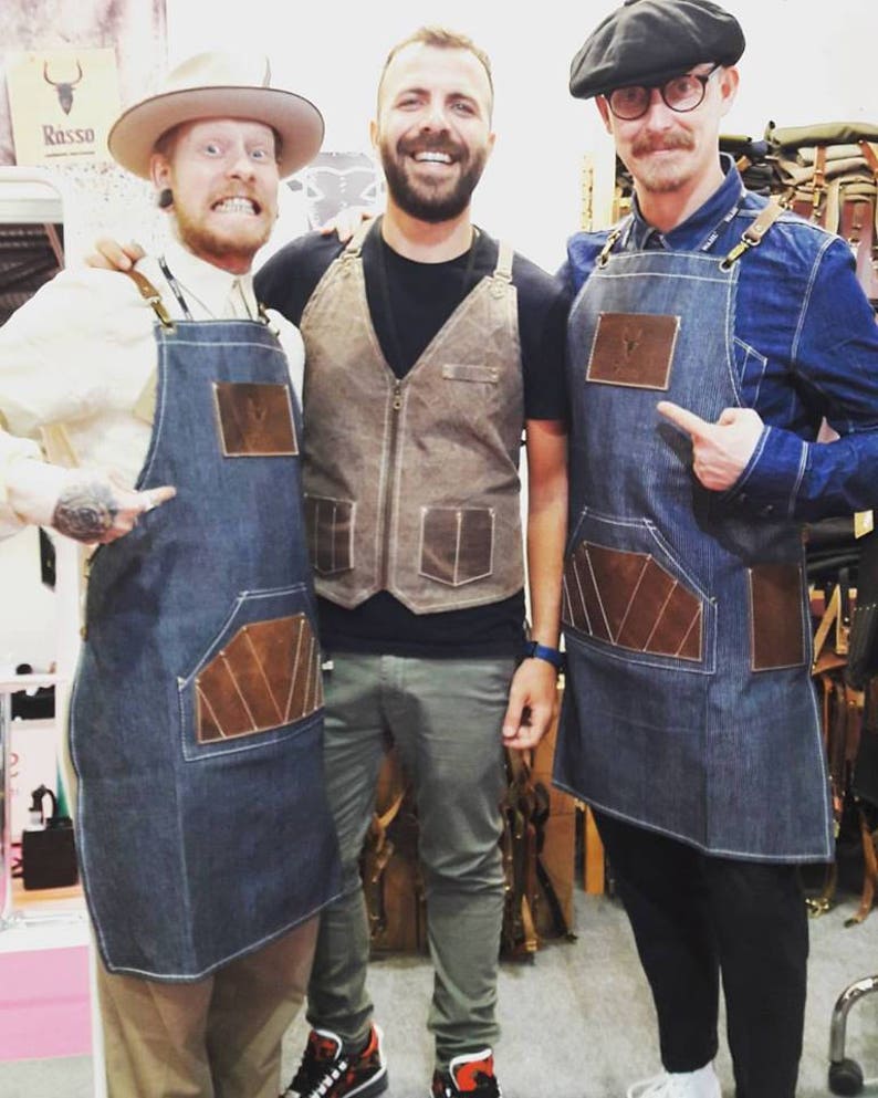 May include: Three people wearing denim aprons with leather accents. The aprons have a large pocket on the front. The person on the left is wearing a cowboy hat. The person in the middle is wearing a brown vest. The person on the right is wearing a black beret and glasses. The text "Rasso" is visible in the background.