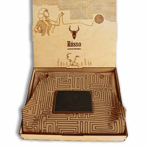 May include: A wooden box with a leather apron inside. The apron has a maze pattern and a leather patch with the word "Rasso" on it. The box lid has a maze pattern and a drawing of a woman holding a ball of yarn.