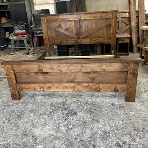 Puede incluir: Un marco de cama de madera con un acabado de tinte marrón. La cabecera presenta un diseño de chevron. El marco de la cama está hecho de madera maciza y tiene un aspecto rústico.