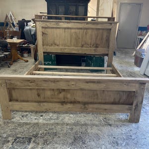 Puede incluir: Un armazón de cama de madera con cabecero y piecero. El armazón de la cama es de madera de color claro y tiene un aspecto rústico e inacabado. El cabecero es rectangular y presenta un diseño de paneles. El piecero también es rectangular y tiene un diseño de paneles similar.