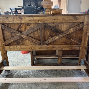 Puede incluir: Un marco de cama rústico de madera con cabecero teñido de marrón oscuro y diseño en forma de X. El marco de la cama está sin terminar, mostrando la veta de la madera. El cabecero es el punto focal, con una estética robusta de estilo rústico.
