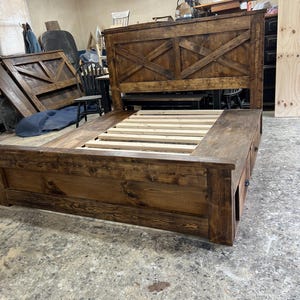 May include: A large, rustic wooden bed frame with a dark brown finish. The headboard features a cross-beam design. The bed frame includes built-in drawers and a slatted base for the mattress. The bed is unfinished and ready for use.