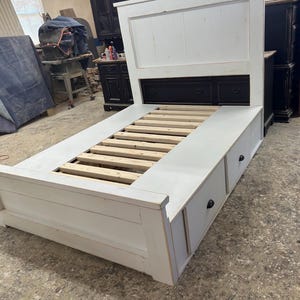 Peut inclure: Un cadre de lit en bois blanc avec une tête de lit et deux tiroirs. La tête de lit est rectangulaire avec un panneau noir en bas. Le cadre de lit a une base à lattes et est peint en blanc.