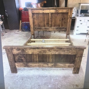 Puede incluir: Una estructura de cama de madera teñida de color marrón oscuro con un cabecero alto y un piecero más bajo con detalles de tablones horizontales. El marco incluye listones de madera clara colocados horizontalmente para el colchón.