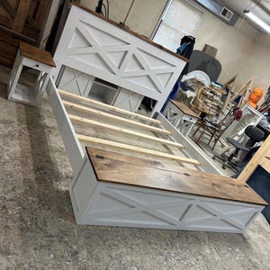 May include: A white and brown wooden bed frame with a matching bedside table. The bed frame features a headboard with an X-shaped design and a wooden top. The bedside table has a similar design. The bed frame is shown without a mattress.