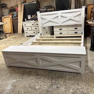 May include: White wooden bed frame with a headboard featuring an X-shaped design. The bed frame has a built-in storage bench at the foot of the bed.