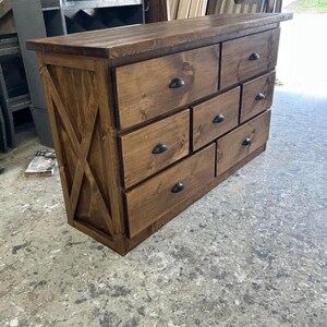 Puede incluir: Una cómoda de madera marrón oscuro con siete cajones y tiradores metálicos negros en forma de semicírculo. El panel lateral muestra un diseño de refuerzo en forma de X. El mueble tiene una superficie superior rectangular y acabado de veta natural.