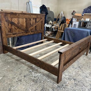 Puede incluir: Una estructura de cama de madera teñida de color marrón oscuro con un cabecero alto que presenta un diseño de refuerzo en X. La estructura incluye un piecero y varias láminas de soporte de madera clara sin acabado a lo ancho.