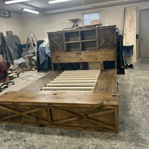 Puede incluir: Un gran marco de cama de madera hecho a mano con una cabecera con estantes de almacenamiento y puertas de armario. El marco de la cama tiene un pie de cama con un diseño en forma de X y un acabado de madera natural.
