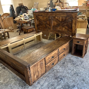 Può includere: Un letto rustico in legno tinto scuro con testiera, pediera e comodino. La testiera presenta un design a porta da fienile. La struttura del letto include cassetti per riporre gli oggetti. Il comodino ha un cassetto e una mensola aperta.