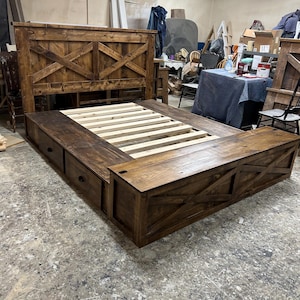 May include: A large, dark-stained wooden bed frame with a headboard featuring an X-shaped design. The bed includes a platform with drawers and a storage bench at the foot. The bed frame is made of solid wood and has a rustic, farmhouse style.