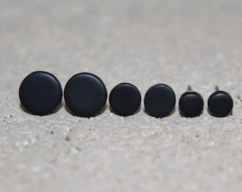 May include: A set of six black, circular stud earrings of varying sizes. The earrings are arranged in a row, showcasing a matte finish. The simple design makes them suitable for everyday wear. The earrings are displayed on a textured, neutral-toned surface.