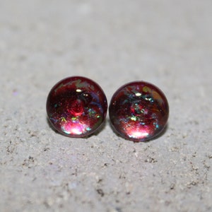 May include: A pair of round, dichroic glass stud earrings. The earrings are a deep burgundy color with iridescent flecks of green, blue, and gold. The studs are set against a textured, gray surface.