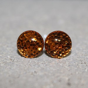 May include: A pair of round stud earrings. Each earring is a clear dome filled with orange glitter. The earrings are set against a neutral gray background. The earrings are a simple, yet eye-catching accessory.