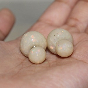 May include: A pair of off-white, double-sided stud earrings. Each earring features a large, round, glittery orb paired with a smaller, round orb. The earrings have a pearlescent sheen and are set against a neutral background.