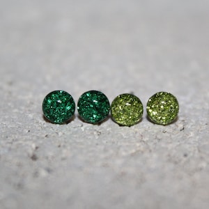 May include: Four round stud earrings with a glittery finish. The earrings are arranged in a row, with two dark green, one medium green, and one light green earring. The earrings are on a gray surface.