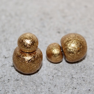 May include: Close-up of gold-colored earrings with a textured, metallic finish. The earrings are in two different styles: one pair features a small sphere atop a larger sphere, and the other pair has a single sphere.