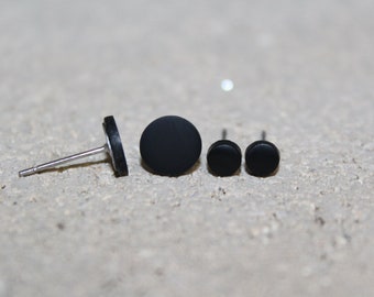 May include: A collection of four black stud earrings in varying sizes. The earrings have a matte finish and are arranged on a light-colored, textured surface. The earring posts are silver.