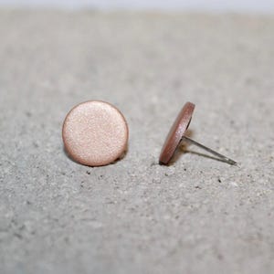 May include: Two round, rose gold stud earrings. One earring is lying flat, while the other is positioned on its side, showing the post. The earrings are on a textured, light gray surface.