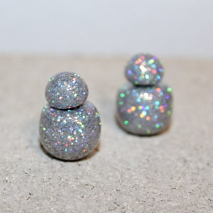 May include: Two silver-colored, double-sided stud earrings. Each earring has a larger, rounded base and a smaller, rounded top. The earrings are covered in iridescent glitter, reflecting multiple colors.