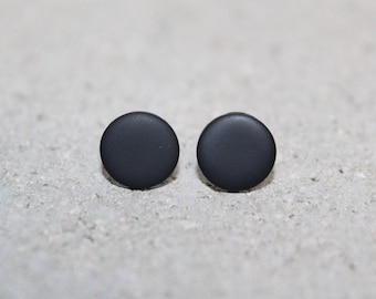 May include: A pair of matte black stud earrings. Each earring is a simple, solid circle. The earrings are displayed on a light gray, textured surface. The earrings are a minimalist design.