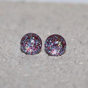 May include: A pair of round stud earrings with a glittery design. The earrings feature a mix of red, blue, and silver glitter, creating a sparkling effect. The studs are set against a neutral gray background.