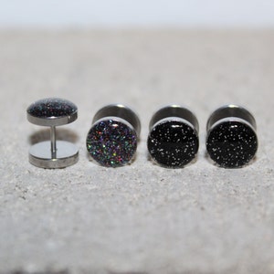 May include: Four stainless steel ear gauges. One is a double-sided plug with a post, and the other three are single-sided plugs. The plugs have a black glitter design, and the other two have a black speckled design.