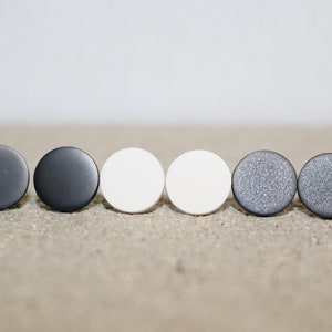May include: A collection of six round stud earrings in various colors. The earrings include shades of gray, white, and a textured silver. The earrings are displayed on a neutral, textured surface.