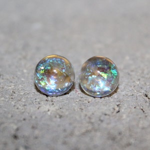May include: Two clear, iridescent glass marbles. The marbles have a spherical shape and display a range of colors, including blue, green, and gold, due to the light refraction. They sit on a gray, textured surface.