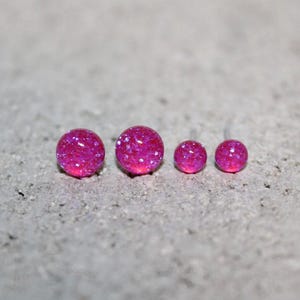 May include: Four round stud earrings in a vibrant hot pink color with a glittery, textured finish. The earrings are arranged in a row, featuring two larger studs on the left and two smaller studs on the right, all with a reflective surface.