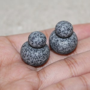 May include: Two gray, speckled, round beads, each with a smaller bead stacked on top. The beads are in a person's open palm. The beads have a speckled pattern of black dots.